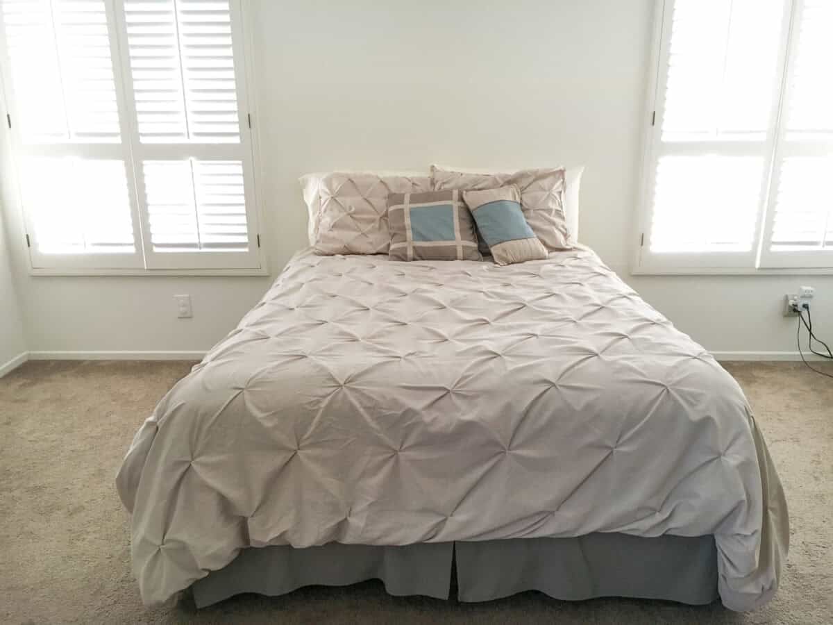 The before - a bland white room with bed in the middle with beige bedspread no headboard