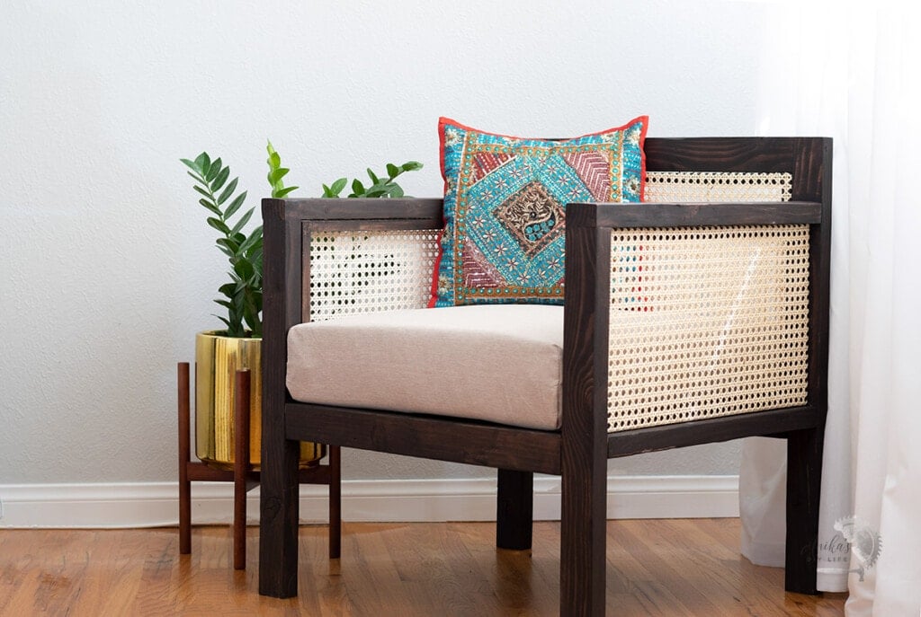Brown chair with cane webbing in arm and back in room with colorful pillow and plant next to it