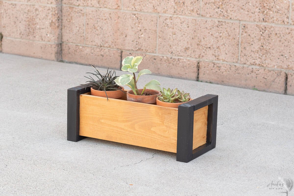 brown planter with black trim with three pots of succulents on a concrete with brick wall in the back.