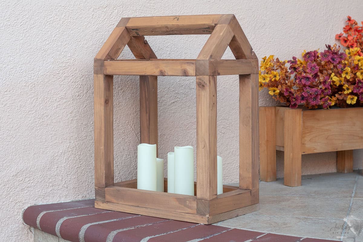 DIY wooden lantern in the shape of a house on porch with candles inside and fall flowers behind.