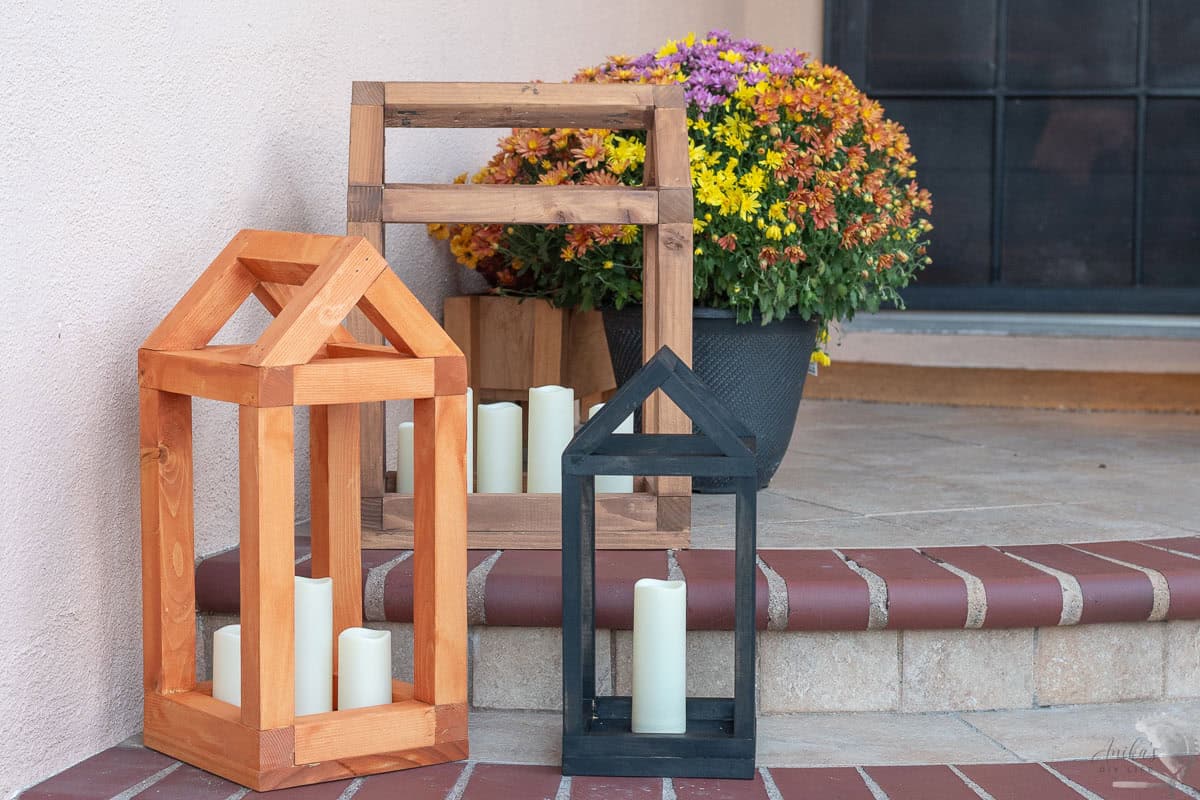 Three house shaped DIY wood lanterns in various sizes sitting on the porch in front of fall flowers.