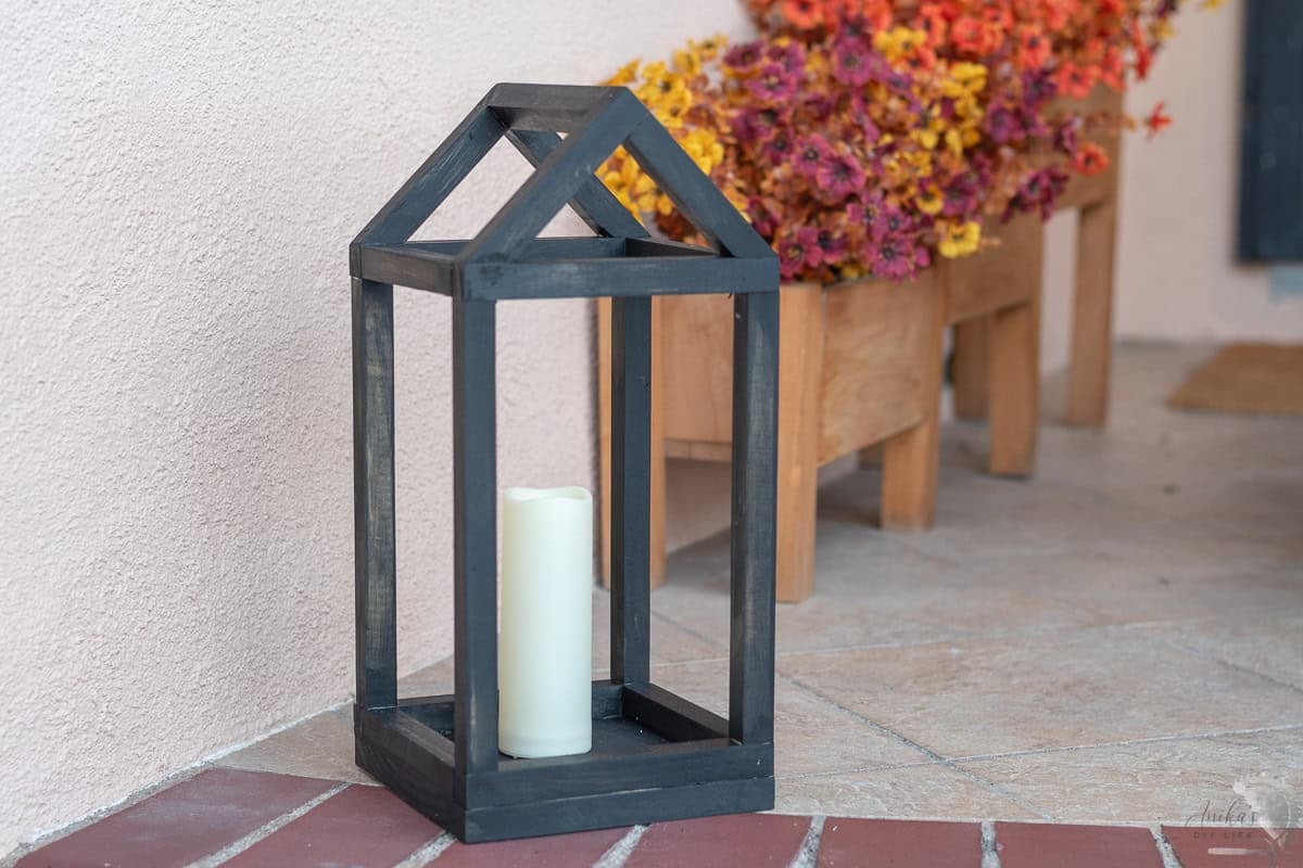 black house shaped DIY wood lantern on porch with candles inside and fall flowers behind
