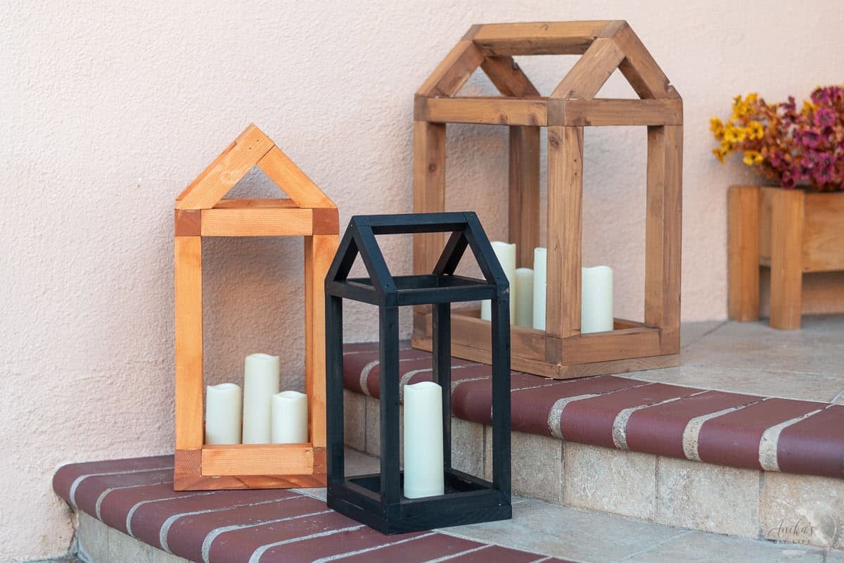 three house shaped DIY wooden lanterns on porch in front of fall flowers.