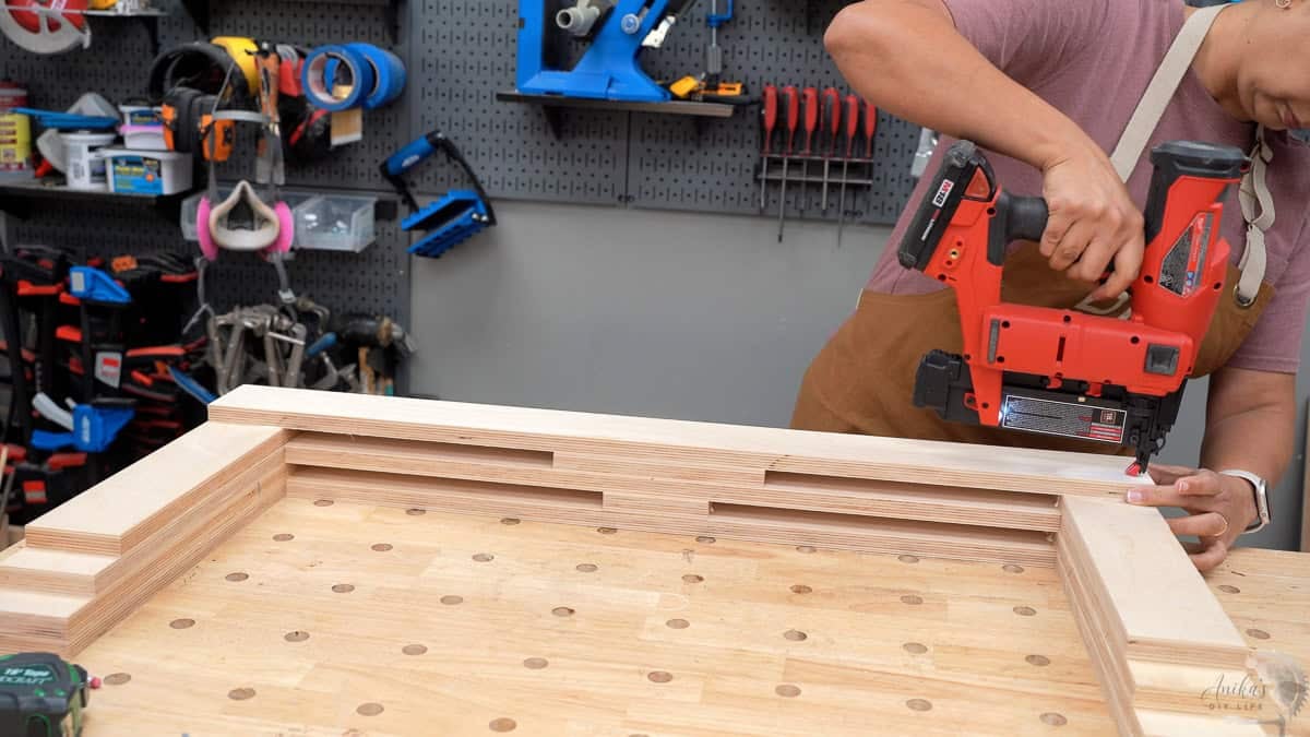 Anika nailing plywood to build the bench in her workshop. There are tools on a pegboard behind. 