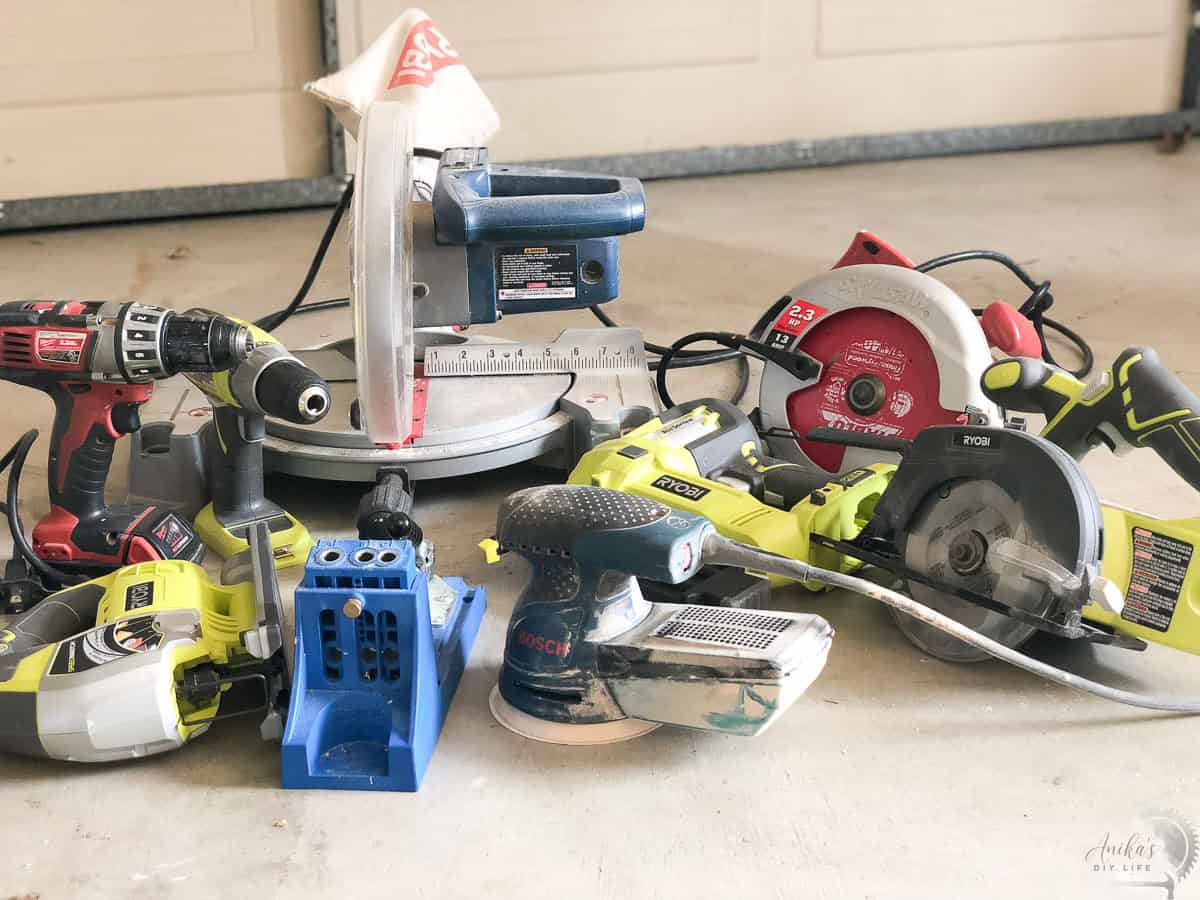 A collection of foundational woodworking tools for beginners, including a miter saw, power drill, and safety gear, arranged on a workshop floor.