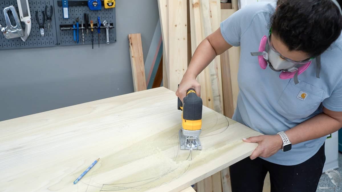 A woodworker wearing safety gear and a respirator while using a yellow jigsaw to cut curved shapes into a wood panel.