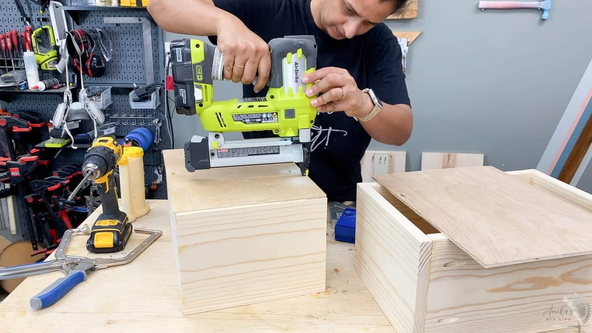 A Ryobi 18-gauge cordless brad nailer being used to assemble a small wooden box on a workshop table.