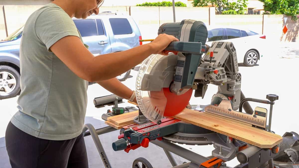 A woman with a pencil in her hand operates a sliding compound miter saw to cut cedar fence pickets for a DIY planter.