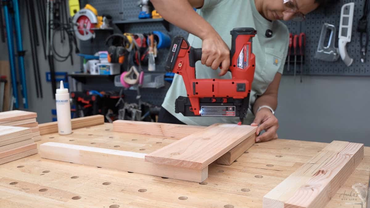 A close-up view of a beginner woodworker using a cordless brad nailer and outdoor glue to assemble the side panels of a square planter.
